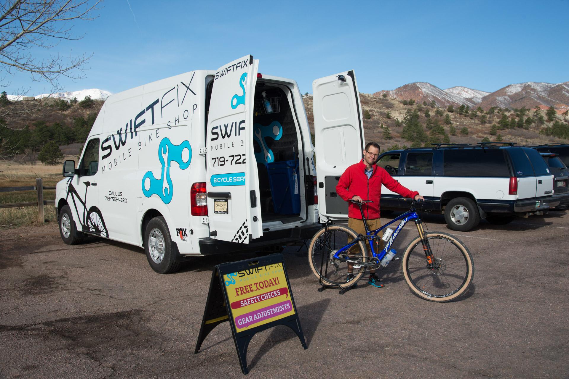 A mobile bike shop van with the name "SWIFTFIX" is parked in an outdoor setting. The van’s back doors are open, revealing tools and equipment inside. A man stands next to a blue mountain bike, holding it while wearing a red jacket. In front of the van, there is a sign advertising free bike safety checks and gear adjustments. The background features trees and hills, set against a clear blue sky.