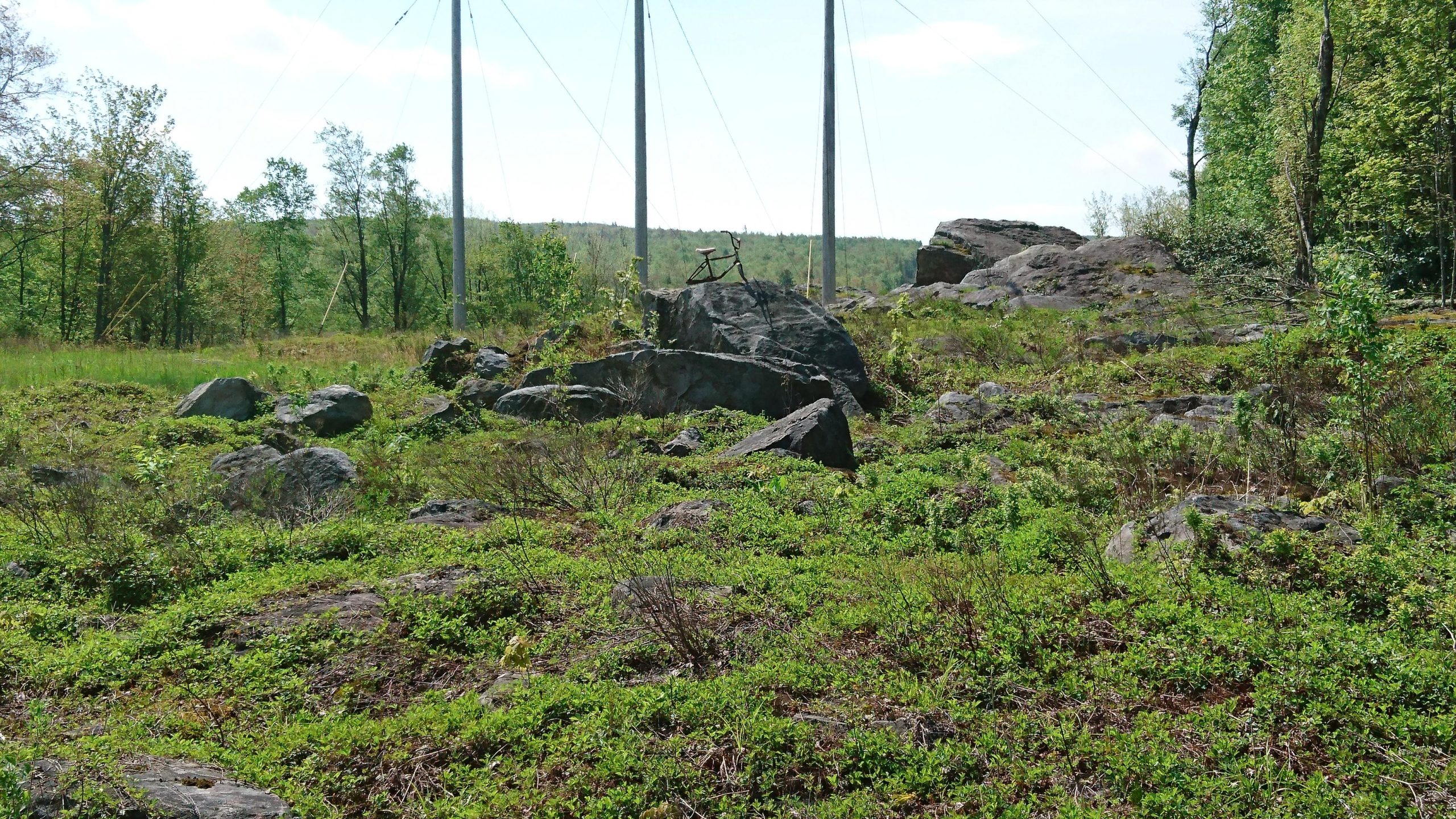 A natural landscape featuring a rocky terrain with patches of green vegetation. In the background, there are several power poles and trees, with a blue sky and wispy clouds visible. A large rock in the foreground has what appears to be a stick or small object resting on it. CVI Trails mountain bike trail.