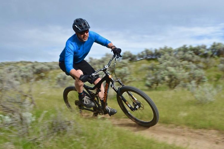 A person wearing a blue long-sleeve shirt, black shorts, and a helmet is riding a mountain bike on a dirt trail surrounded by green grass and sparse shrubs. The biker is leaning into a turn, showcasing an action shot in a natural outdoor setting. The sky is partly cloudy, indicating an overcast day.