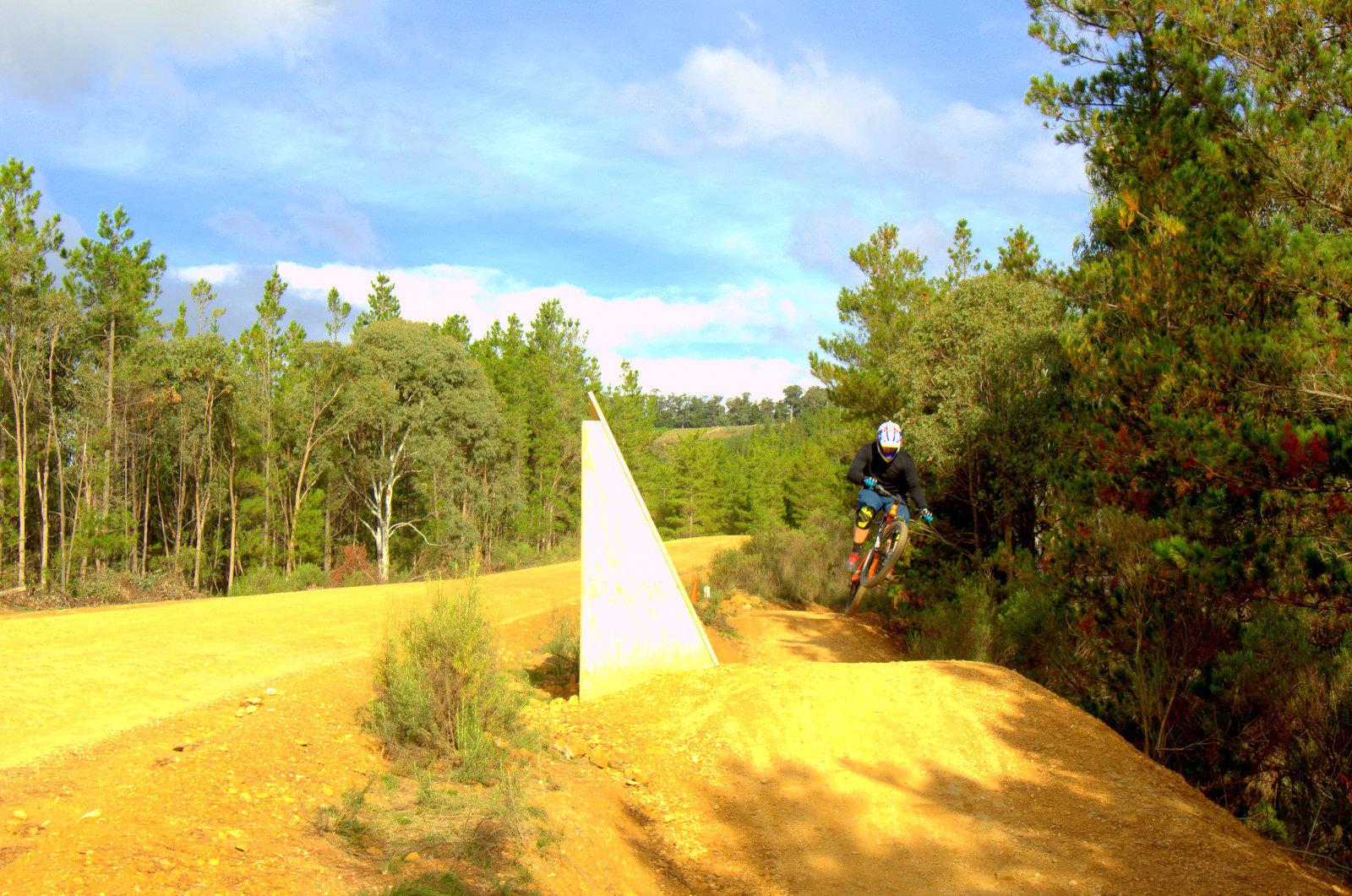 A mountain biker jumping off a dirt ramp near an angled wooden feature, surrounded by trees and a clear blue sky. The scene captures the excitement of outdoor biking in a natural setting. Hero Trail mountain bike trail.