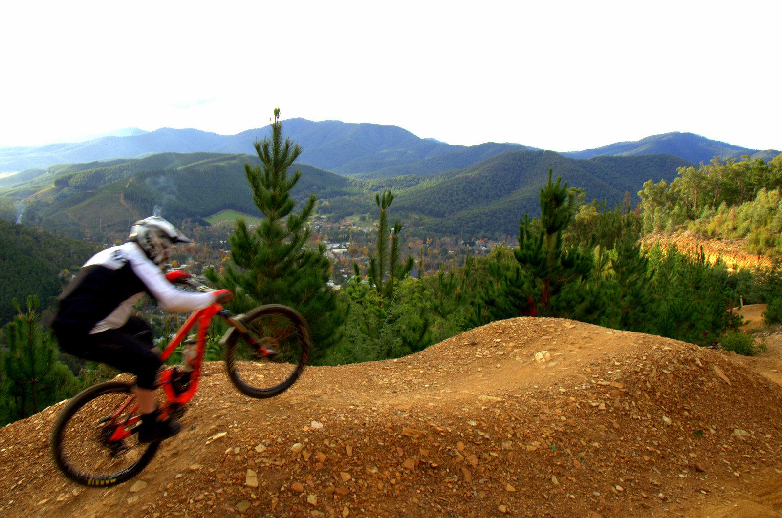 A mountain biker performing a jump on a dirt trail with a scenic view of rolling hills and a small town in the distance, surrounded by green trees. The sky is partly cloudy. Hero Trail mountain bike trail.