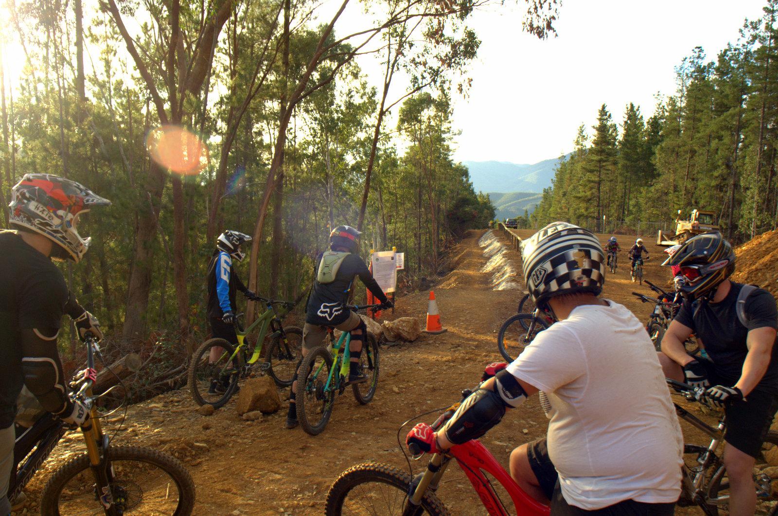 A group of mountain bikers in helmets and protective gear pause on a dirt trail surrounded by trees. Some bikers sit on their bikes while others stand, observing a sign ahead. The scene is bathed in warm sunlight with mountains visible in the distance. Hero Trail mountain bike trail.