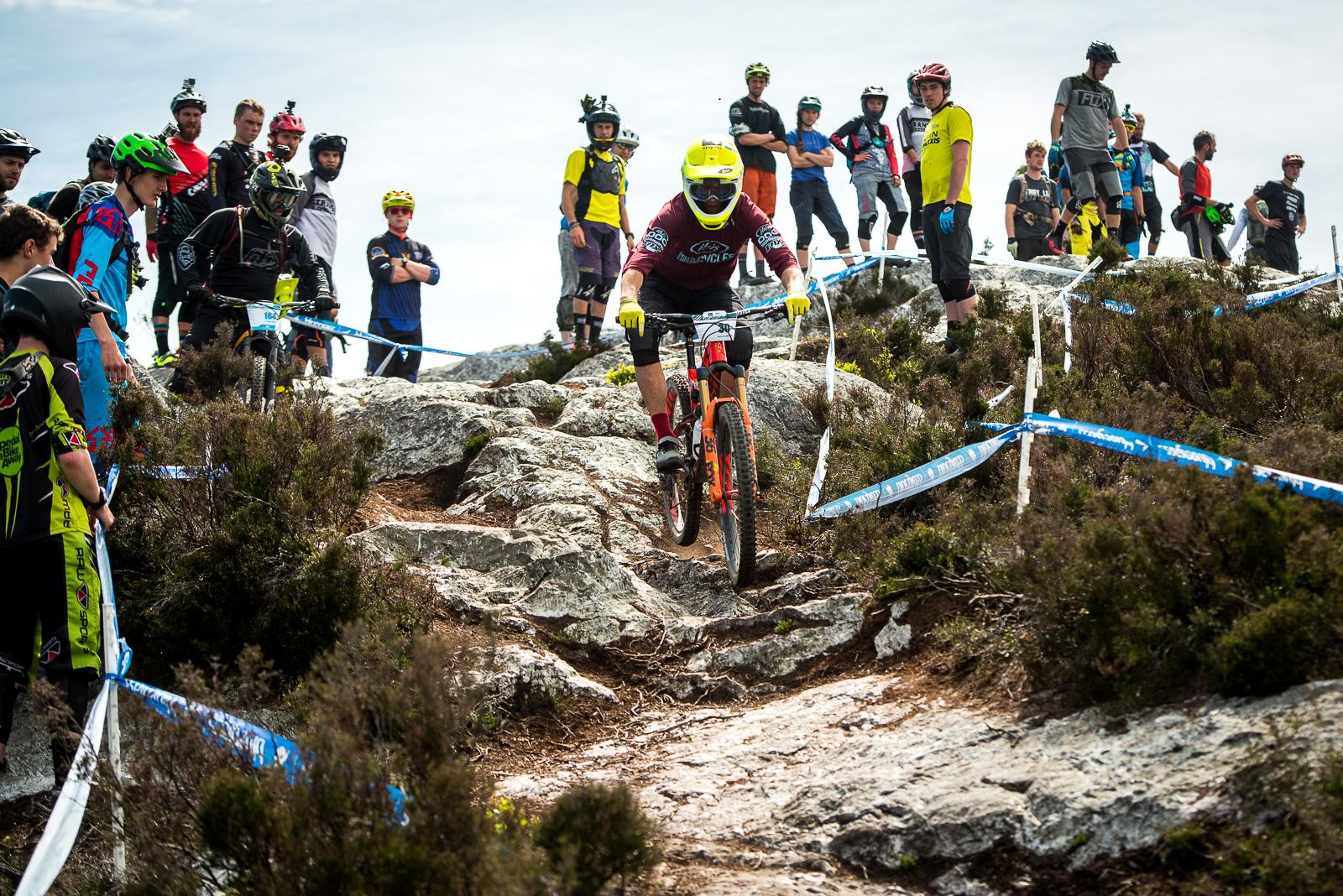 Ibis Mojo HD4: A mountain biker navigates a rocky path during a downhill race, while a crowd of spectators watches on the sidelines. The scene is set in an outdoor environment with scattered rocks and greenery, and safety tape marking the course. Various riders and onlookers, dressed in colorful biking gear and helmets, create a lively atmosphere.