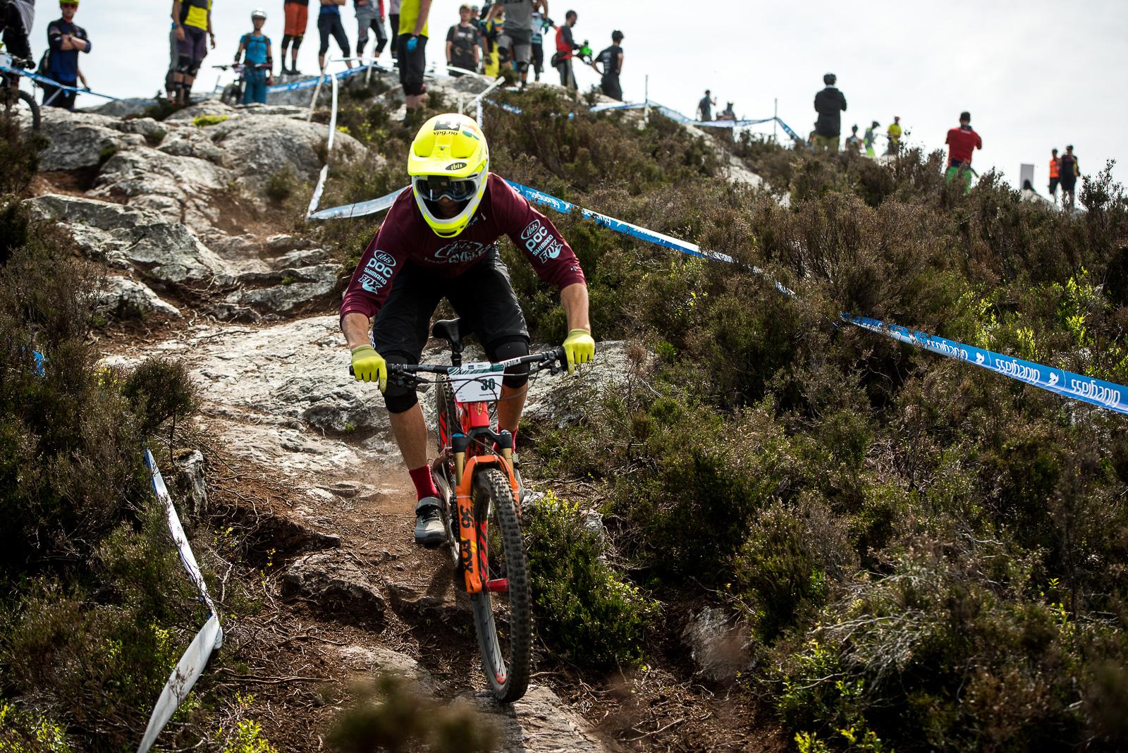 Ibis Mojo HD: A mountain bike racer navigates a rocky downhill trail, leaning into a turn. The rider wears a bright yellow helmet and protective gear, with a focus on maintaining balance. In the background, a crowd of spectators watches from various vantage points along the trail, creating an atmosphere of excitement during the race.