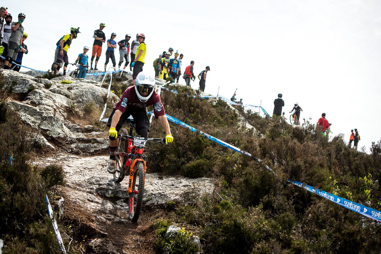 Ibis Mojo HD: A mountain biker navigates a rocky trail during a downhill biking event, while a crowd of spectators watches from an elevated position. The scene is set in a natural landscape with sparse vegetation and a clear sky. The rider is wearing protective gear, including a helmet and gloves, highlighting the intensity of the sport. Brightly colored banners and markings are visible along the trail.