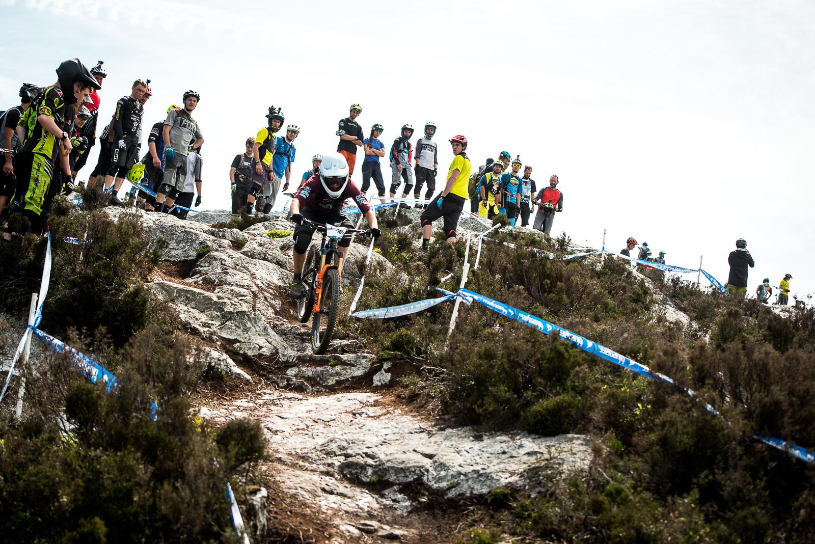 Ibis Mojo HD: A mountain biker descends a rocky trail while a crowd of spectators watches nearby. The scene captures the excitement of a downhill biking event, with the biker in protective gear maneuvering through the rugged terrain. The spectators, dressed in various cycling attire, are positioned along the trail's edge, adding to the atmosphere of the event.