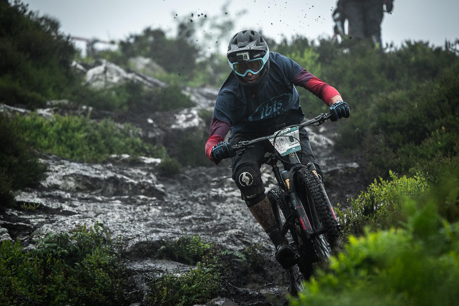 Ibis Mojo HD4: A mountain biker navigating a muddy, rocky trail during rainy weather, wearing a helmet and protective gear. The surrounding landscape features lush greenery and rocks, with splashes of mud in the air from the bike's tires.