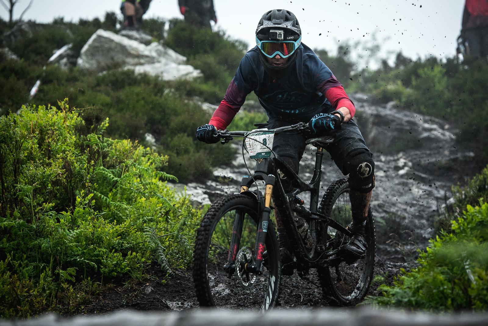 Ibis Mojo HD: A mountain biker navigates a muddy trail surrounded by greenery, wearing protective gear and goggles, with rain falling and mud splattering from the bike tires.