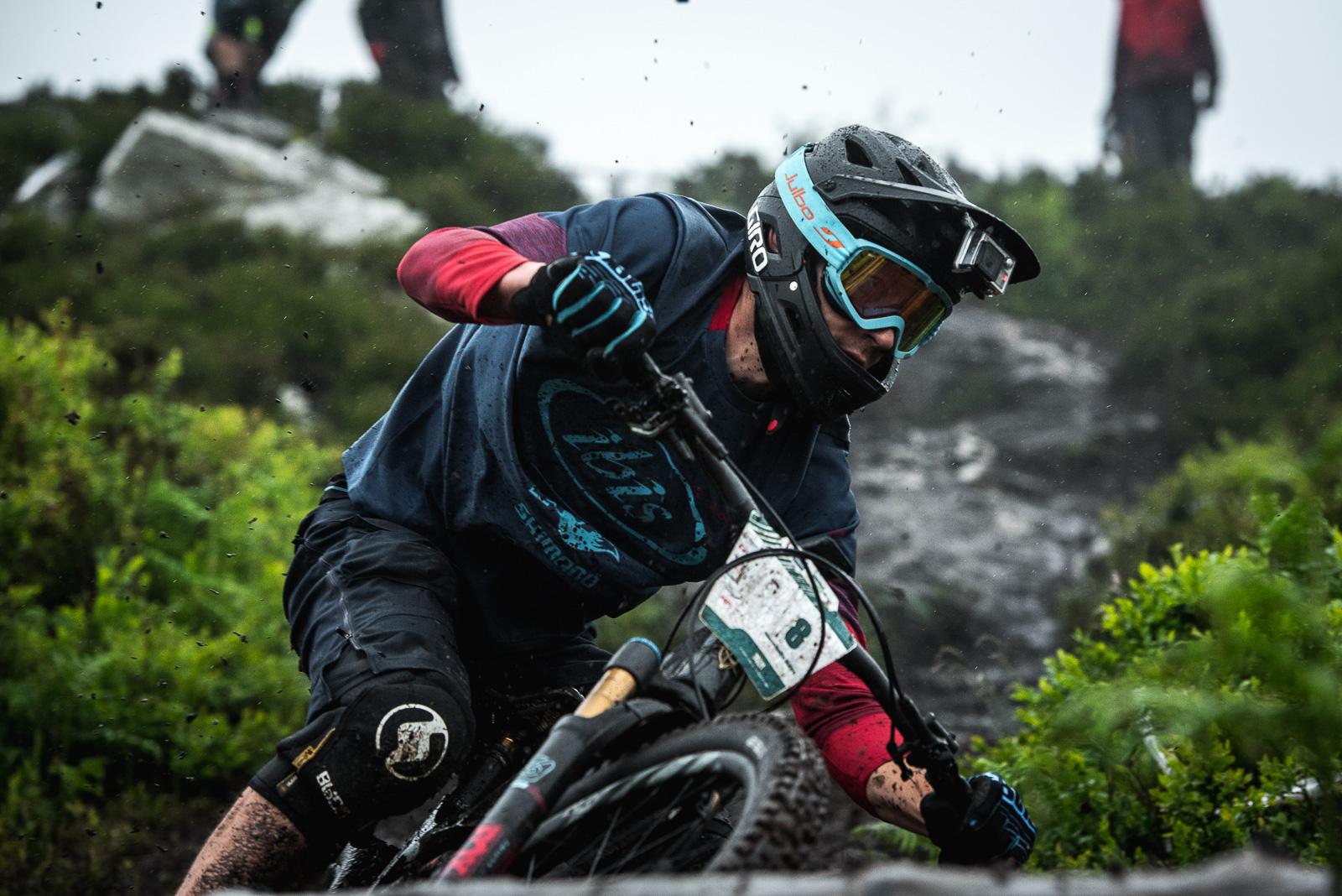 Ibis Mojo HD: A mountain biker leans into a curve on a muddy trail, wearing a helmet and goggles, while rain splashes around him. The background features lush greenery and rocky terrain, with a blurred figure in the distance.