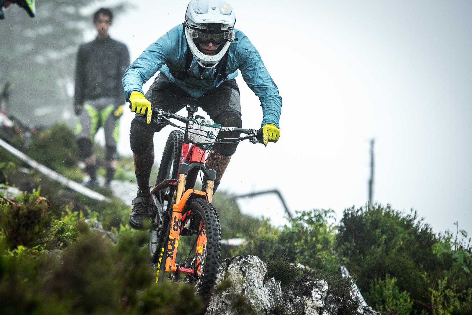 Ibis Mojo HD: A mountain biker in a blue jacket and yellow gloves navigates rocky terrain while riding downhill in rainy conditions. Another rider can be seen in the background, highlighting the challenging outdoor environment.