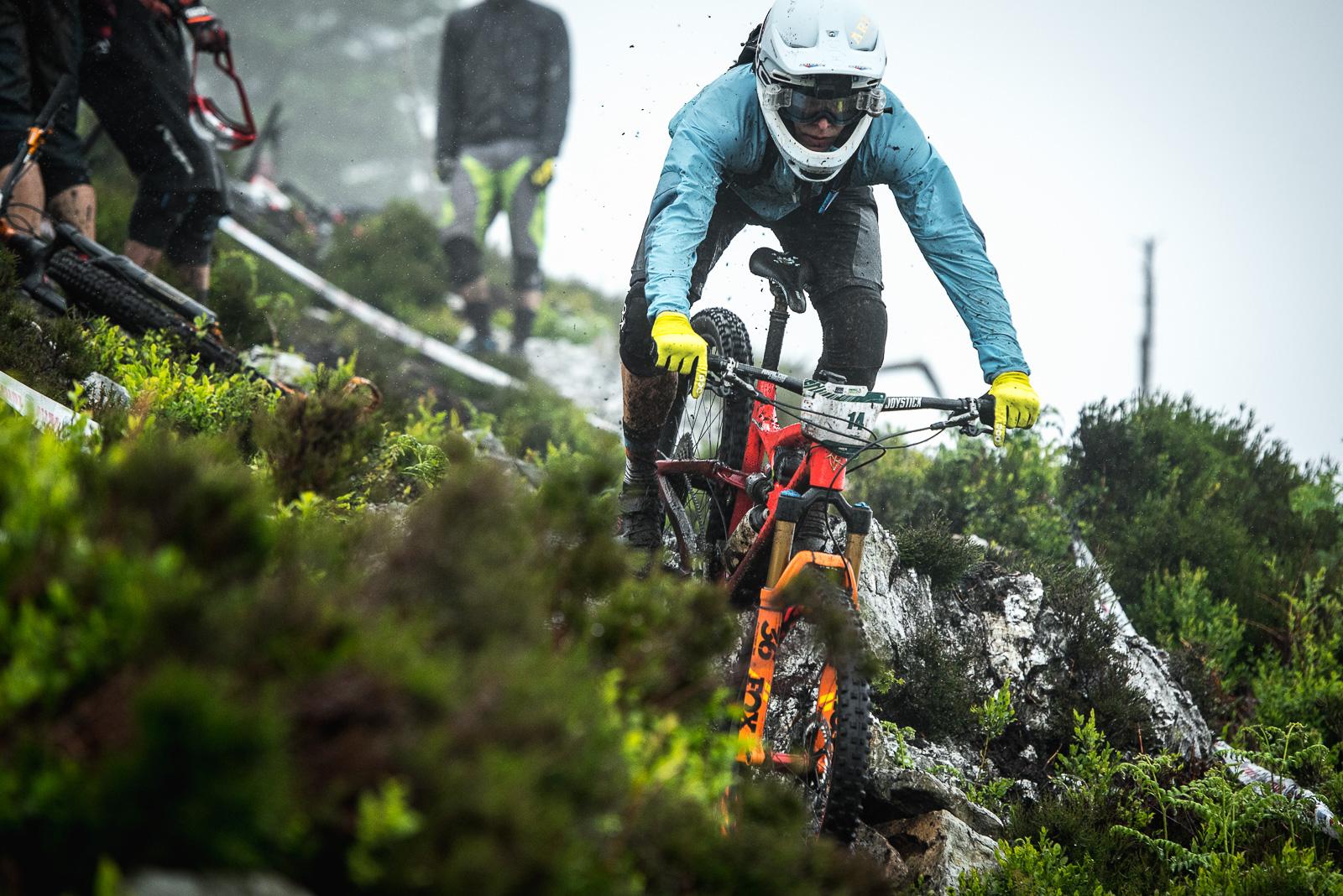 Ibis Mojo HD: A mountain biker navigates a rocky, wet trail in a misty environment, wearing a helmet, goggles, and protective gear. The rider is focused on maneuvering the bike, which features a bright red and orange frame, while splashes of mud and water are kicked up from the tires. In the background, other cyclists are partially visible, surrounded by lush greenery. The atmosphere is dynamic and intense, reflecting the challenges of off-road cycling in adverse weather conditions.