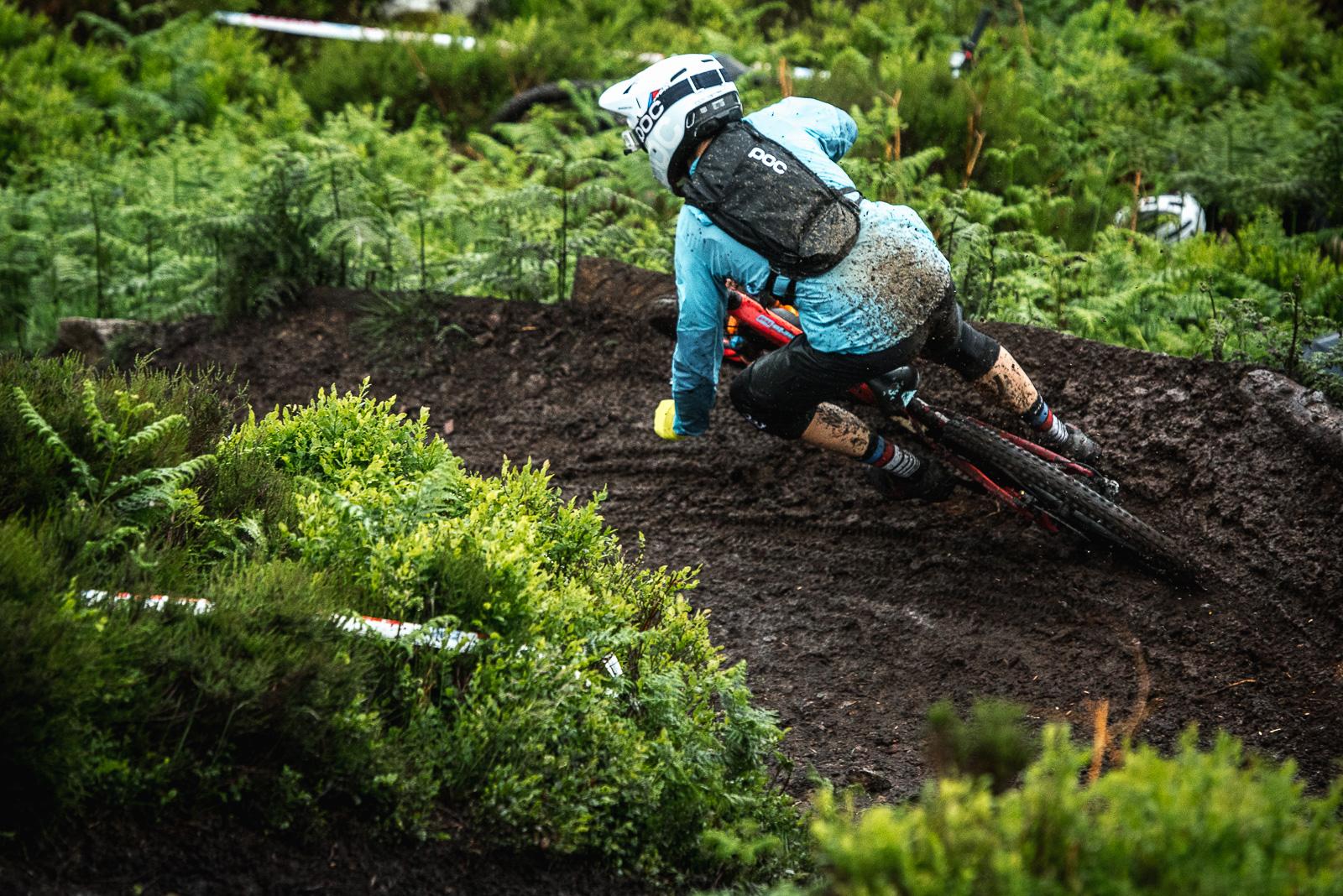 Ibis Mojo HD4: A mountain biker dressed in a blue shirt and black shorts leans into a turn on a muddy trail surrounded by lush greenery. The rider is covered in mud, indicating the challenging conditions of the terrain.