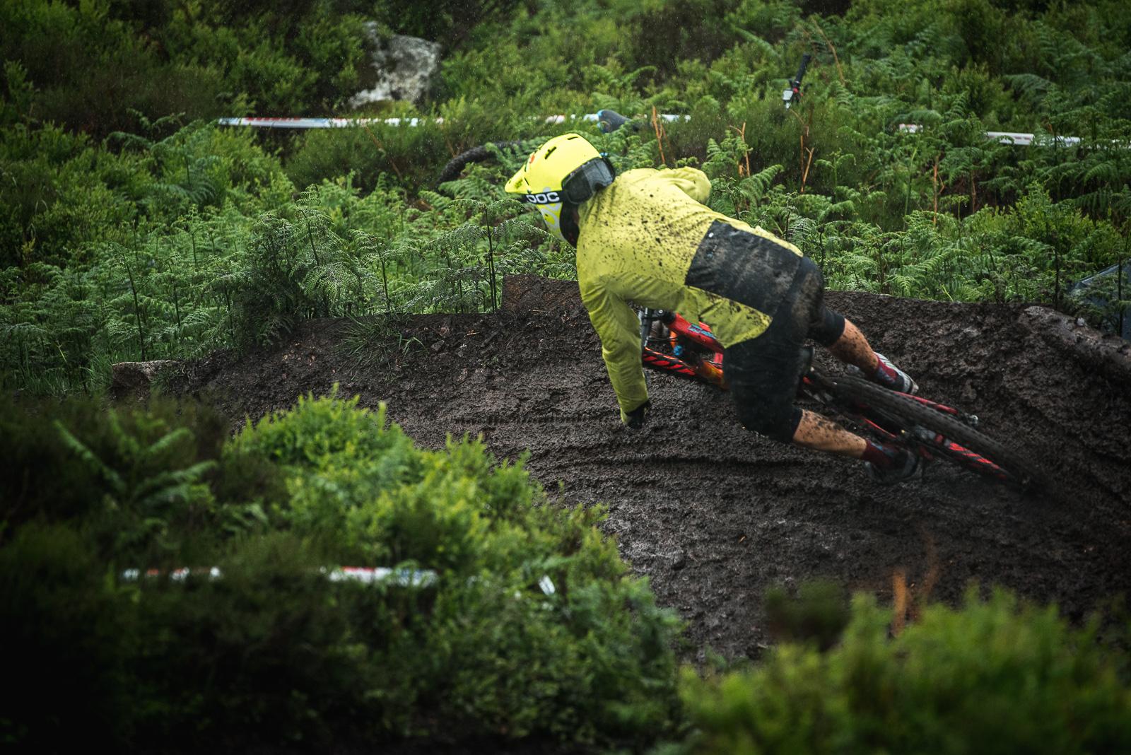 Ibis Mojo HD4: A mountain biker in a bright yellow rain jacket and helmet navigates a muddy turn on a trail surrounded by lush greenery. The rider is leaning into the curve, with mud splattered on their clothing, capturing the intensity of riding in wet conditions. Rain is visible in the atmosphere, adding to the rugged outdoor experience.