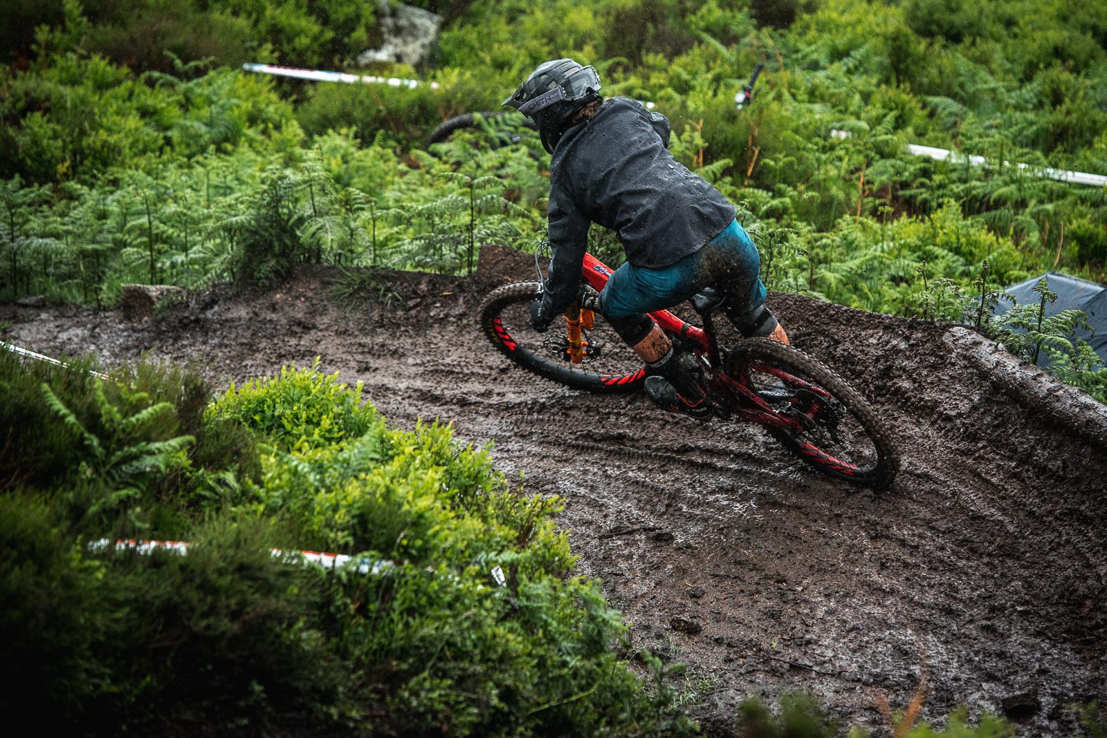 Ibis Mojo HD: A mountain biker maneuvering through a muddy trail, leaning into a curve surrounded by lush greenery. The rider is dressed in a black jacket and blue shorts, with protective gear, illustrating an adventurous scene in wet conditions.