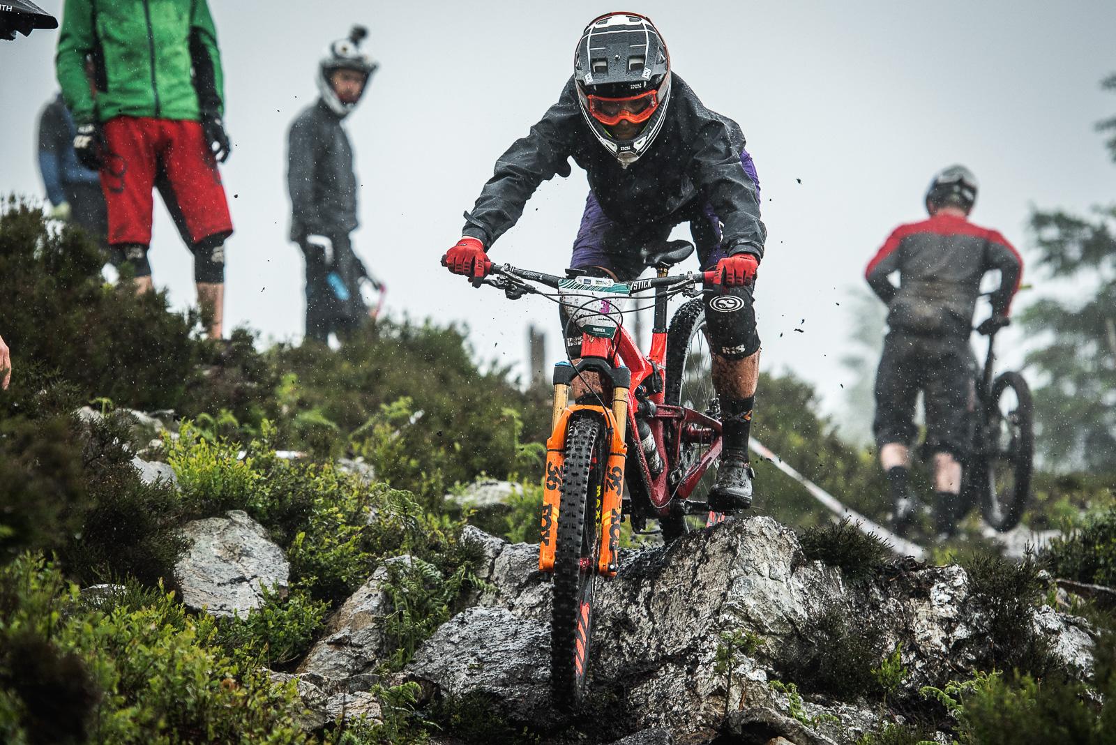 Ibis Mojo HD4: A mountain biker skillfully navigates over rocky terrain in rainy weather, wearing a helmet and protective gear. Several other cyclists are visible in the background, some observing and others preparing to ride. The scene captures the excitement of downhill biking in a natural outdoor setting, with droplets of water and mud splashing around.