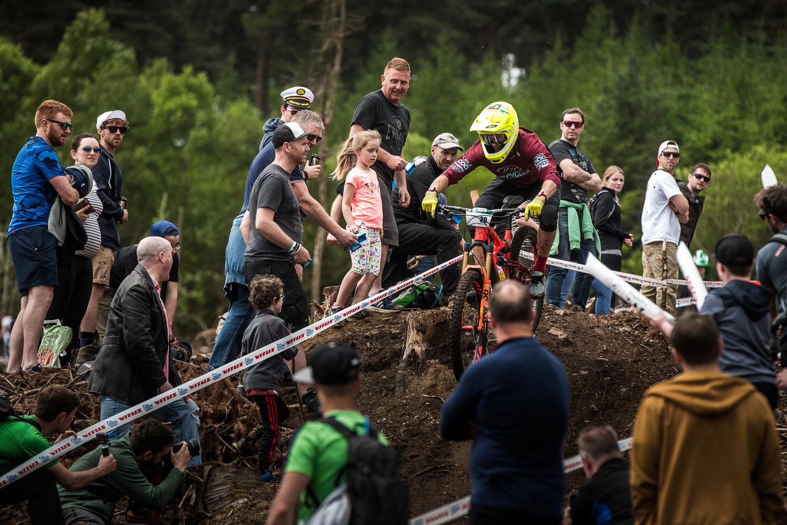 Ibis Mojo HD: A mountain biker in a yellow helmet and protective gear navigates a steep, dirt hill, surrounded by a cheering crowd of spectators, including adults and children. The scene captures the excitement of a mountain biking event in a wooded area, with trees in the background and a racing tape marking the course.