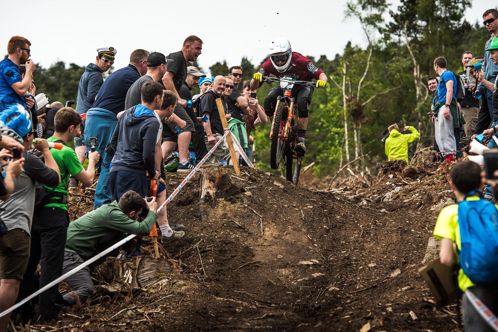 Ibis Mojo HD4: A mountain biker in action, leaping off a dirt jump surrounded by an enthusiastic crowd. Spectators, some holding cameras and phones, cheer from the sides as trees and a forested backdrop are visible in the background. The scene captures the excitement of a downhill mountain biking event, showcasing the thrill and skill of the rider.