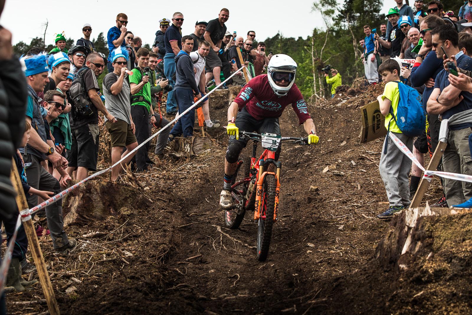 Ibis Mojo HD: A mountain biker in a helmet and protective gear navigates a steep, muddy trail surrounded by a crowd of spectators. The biker is focused and in motion, while onlookers, including children and adults, cheer him on from both sides of the track. The scene captures the excitement of downhill biking in an outdoor setting.