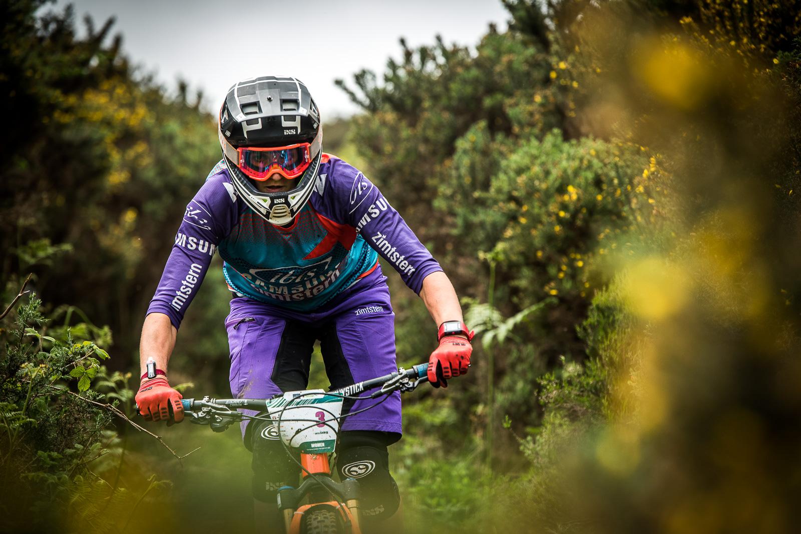 Ibis Mojo HD: A mountain biker in protective gear, including a helmet and gloves, rides through a lush, green trail surrounded by bushy vegetation. The rider is focused and leaning forward on a colorful bike as they navigate the terrain.