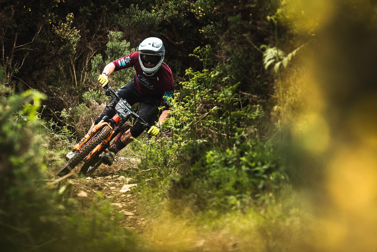 Ibis Mojo HD4: A mountain biker in protective gear leans into a curve on a dirt trail surrounded by lush greenery. The bike features orange accents, and the rider is focused on navigating the terrain. Sunlight filters through the foliage, highlighting the dynamic movement and adventurous spirit of the scene.