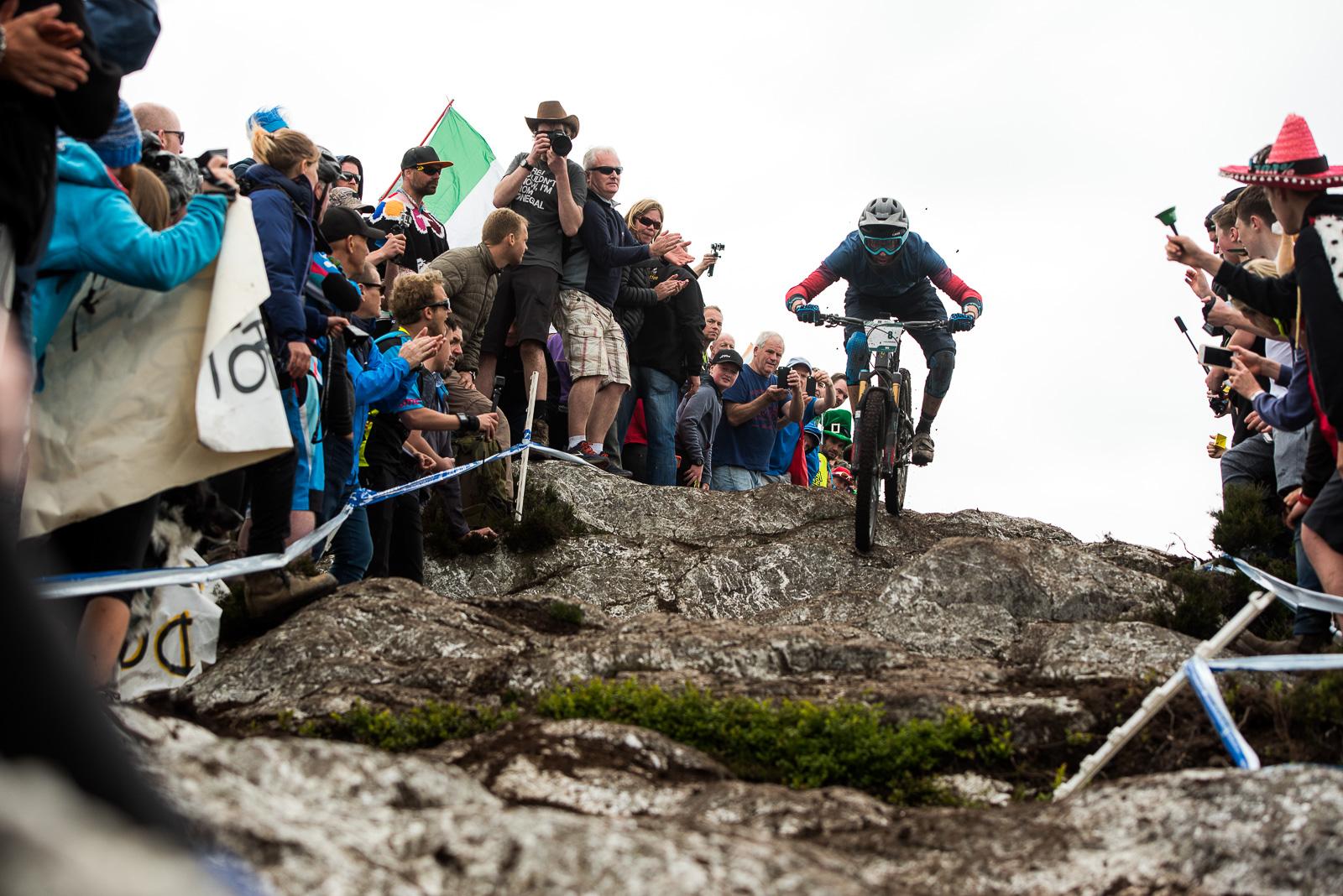 Ibis Mojo HD4: A mountain biker navigates a rocky downhill course while being cheered on by an enthusiastic crowd. The scene captures spectators on both sides of the path, many holding cameras and banners, as they encourage the rider. The cloudy sky sets the backdrop for this exciting outdoor event.