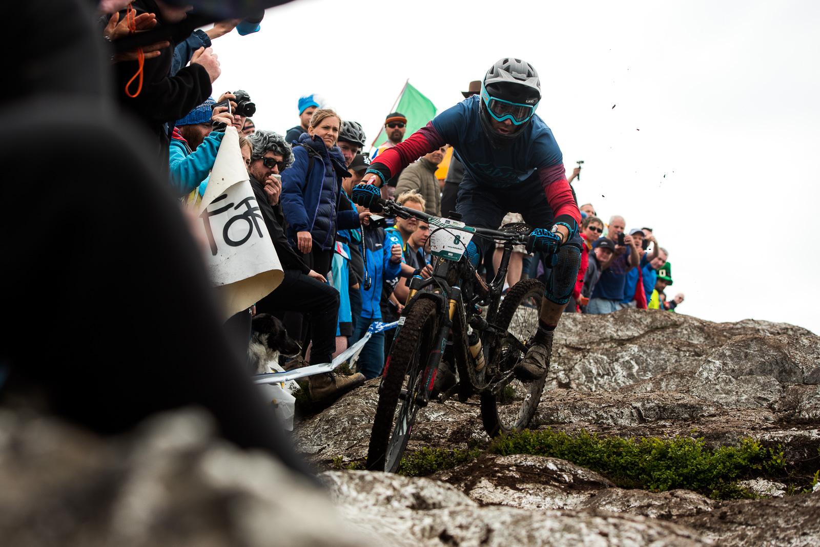 Ibis Mojo HD4: A mountain biker navigates a rocky terrain during a competitive event, with a cheering crowd of spectators in the background. The rider is focused and wearing protective gear, with a mix of colors in their outfit. Some spectators are holding banners and capturing the moment with their cameras. The environment appears rugged, hinting at an exciting mountain biking challenge.