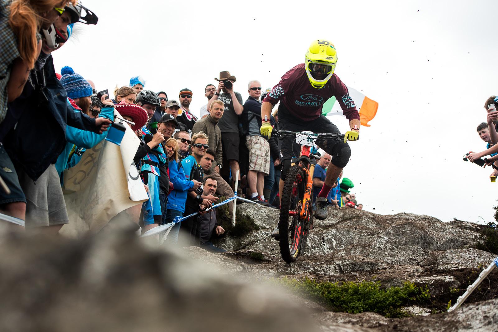 Ibis Mojo HD4: A mountain biker in a yellow helmet and maroon jersey races down a rocky slope, surrounded by a cheering crowd. Spectators, dressed in colorful attire, hold up cameras and banners, capturing the action. An Irish flag can be seen among the crowd, emphasizing the energy and excitement of the event. Dust and debris fly as the biker navigates the challenging terrain.