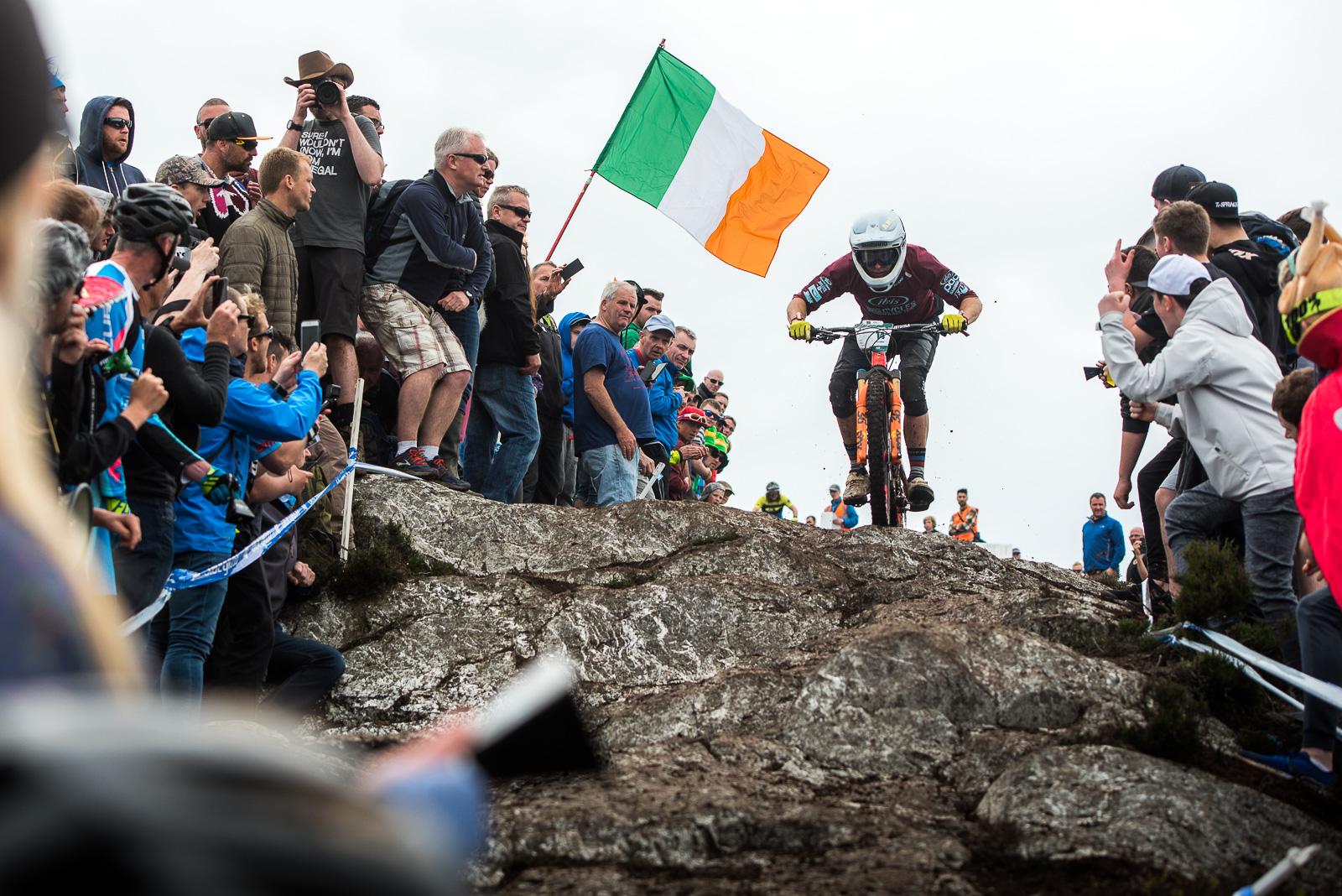Ibis Mojo HD4: A downhill mountain biker navigates a rocky terrain while a crowd watches enthusiastically, with an Irish flag visible in the background. The scene captures the excitement of an outdoor racing event, showcasing both the rider's skill and the supportive atmosphere of the spectators.