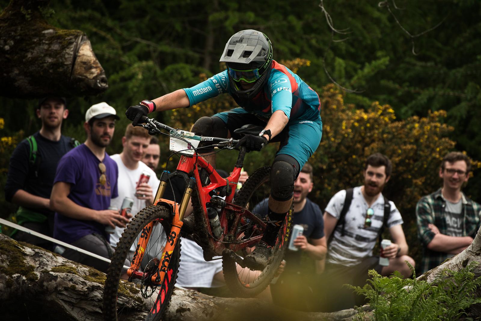 Ibis Mojo HD4: A mountain biker in a teal and red outfit jumps over a log in a forested area, surrounded by a group of onlookers enjoying the event. The biker wears a helmet and goggles, while the spectators react with excitement, some holding drinks. The scene captures the thrill of mountain biking in a natural setting.