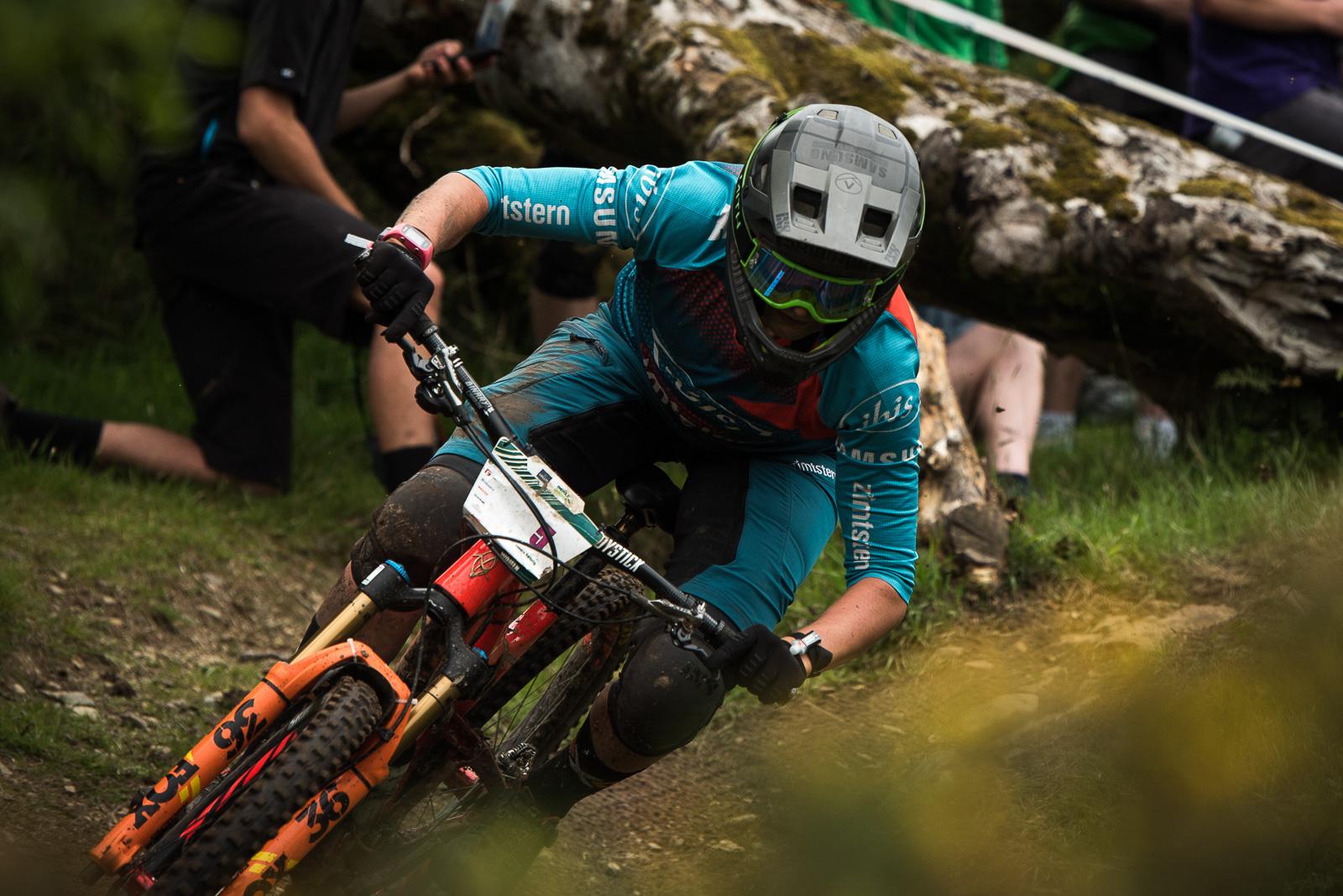 Ibis Mojo HD4: A mountain biker in protective gear leans into a sharp turn on a dirt trail, showcasing skill and focus. A log lies on the ground nearby, indicating a rugged outdoor setting. Spectators are partially visible in the background, adding to the atmosphere of the competitive event. The biker's vibrant blue and orange outfit contrasts with the earthy tones of the trail.