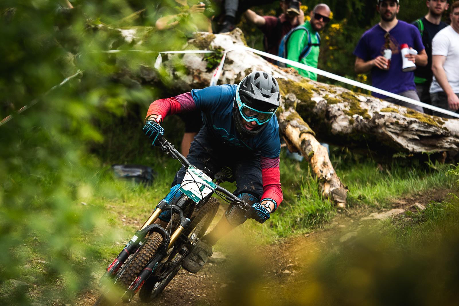 Ibis Mojo HD4: A mountain biker in protective gear leans into a turn on a dirt trail surrounded by greenery. In the background, a group of spectators watches from the side, with a fallen log and some bright foliage visible. The biker wears a blue and red jersey, gloves, and a helmet with goggles, showcasing a dynamic racing moment in an outdoor mountain biking event.