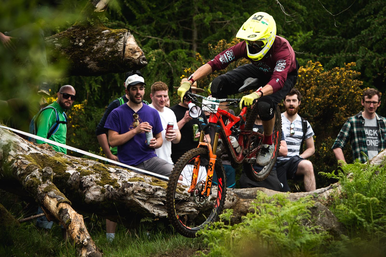 Ibis Mojo HD4: A mountain biker wearing a bright yellow helmet and gloves jumps over a fallen log, while a group of spectators watches and cheers in the background. The scene is set in a lush green environment, showcasing enthusiasts enjoying an outdoor biking event.