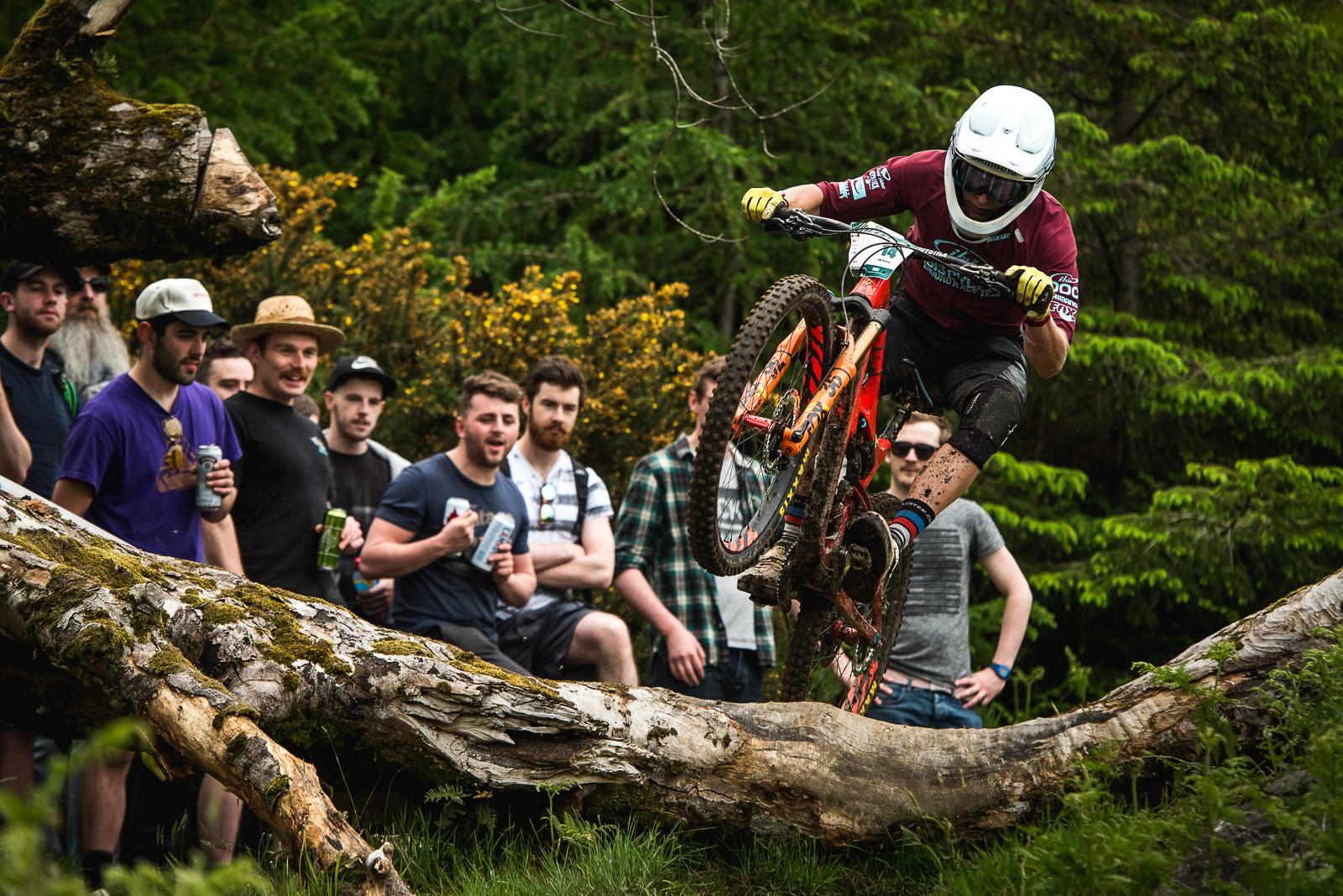 Ibis Mojo HD4: A mountain biker performs a jump over a fallen log in a forested area, while a crowd of spectators, some holding drinks, watches in excitement. The scene captures the thrill of outdoor biking and the enthusiasm of the onlookers.
