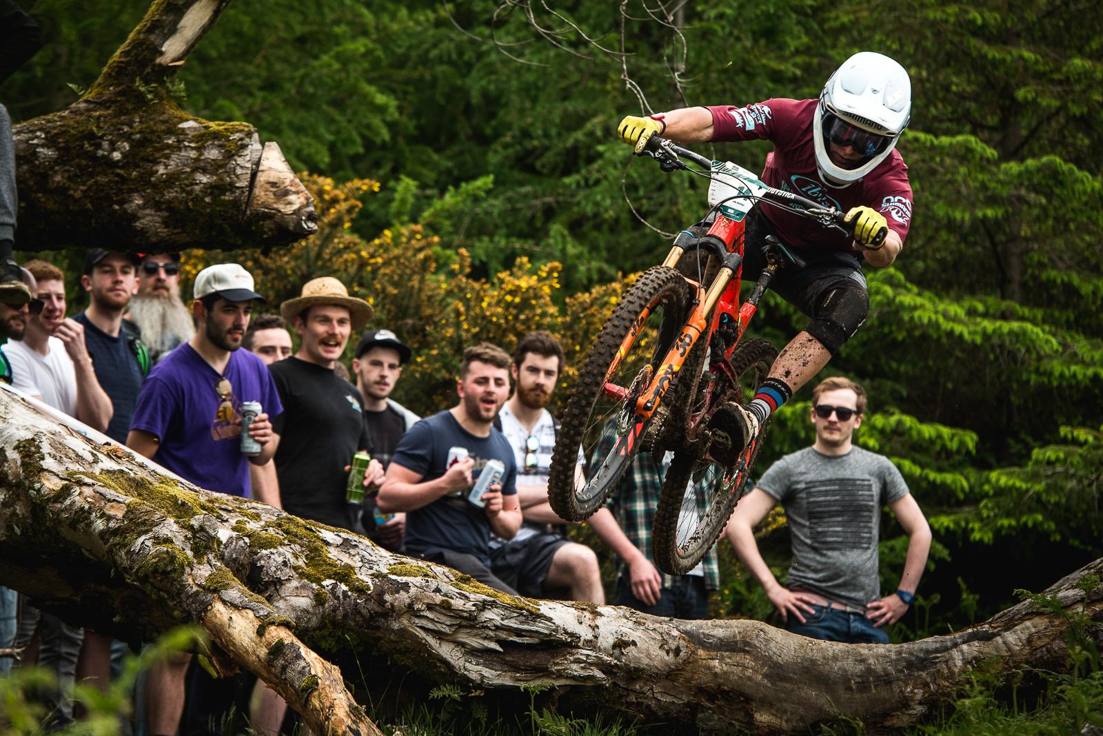 Ibis Mojo HD4: A mountain biker in a helmet and protective gear jumps over a fallen log while performing tricks. A group of enthusiastic spectators watches in the background, some holding drinks, surrounded by lush greenery and flowers.