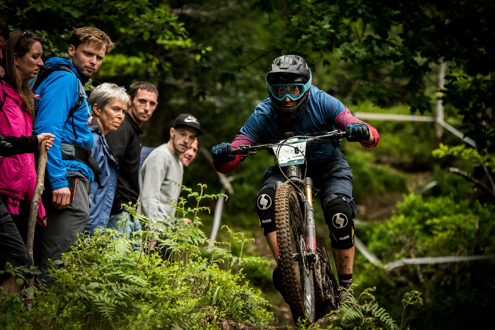 Ibis Mojo HD4: A mountain biker wearing protective gear navigates a challenging dirt trail while several spectators watch intently from the side, surrounded by greenery.