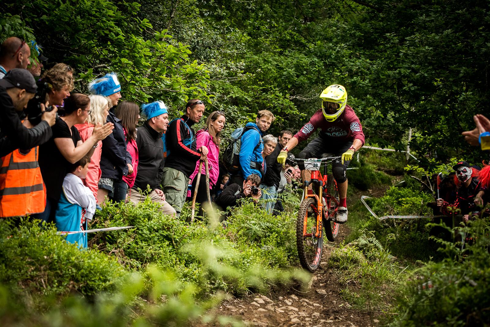 Ibis Mojo HD4: A mountain biker in a yellow helmet and maroon jersey navigates a rocky trail surrounded by a cheering crowd. Spectators are positioned along the path, some wearing colorful accessories, capturing the action with cameras. Lush greenery frames the scene, creating a lively atmosphere during a biking event.