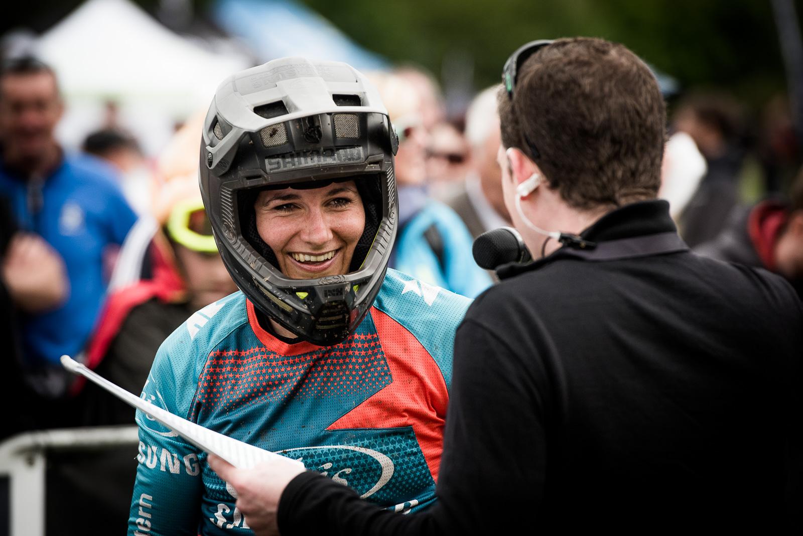 Ibis Mojo HD4: A cyclist wearing a helmet and colorful jersey smiles while interacting with a man holding a microphone and notes. The background features a crowd and outdoor event setting, hinting at a biking competition or festival.