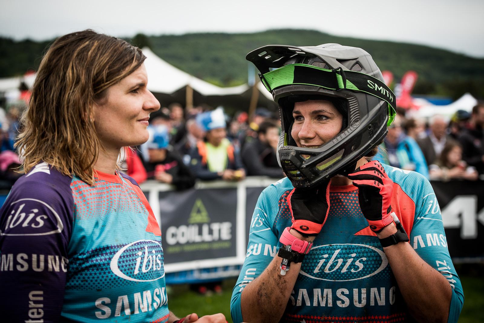 Ibis Mojo HD4: Two female mountain bikers are engaged in conversation at an outdoor event. One is wearing a helmet, while both are dressed in brightly colored cycling jerseys with logos. The background features a crowd and tents, indicating a lively atmosphere.