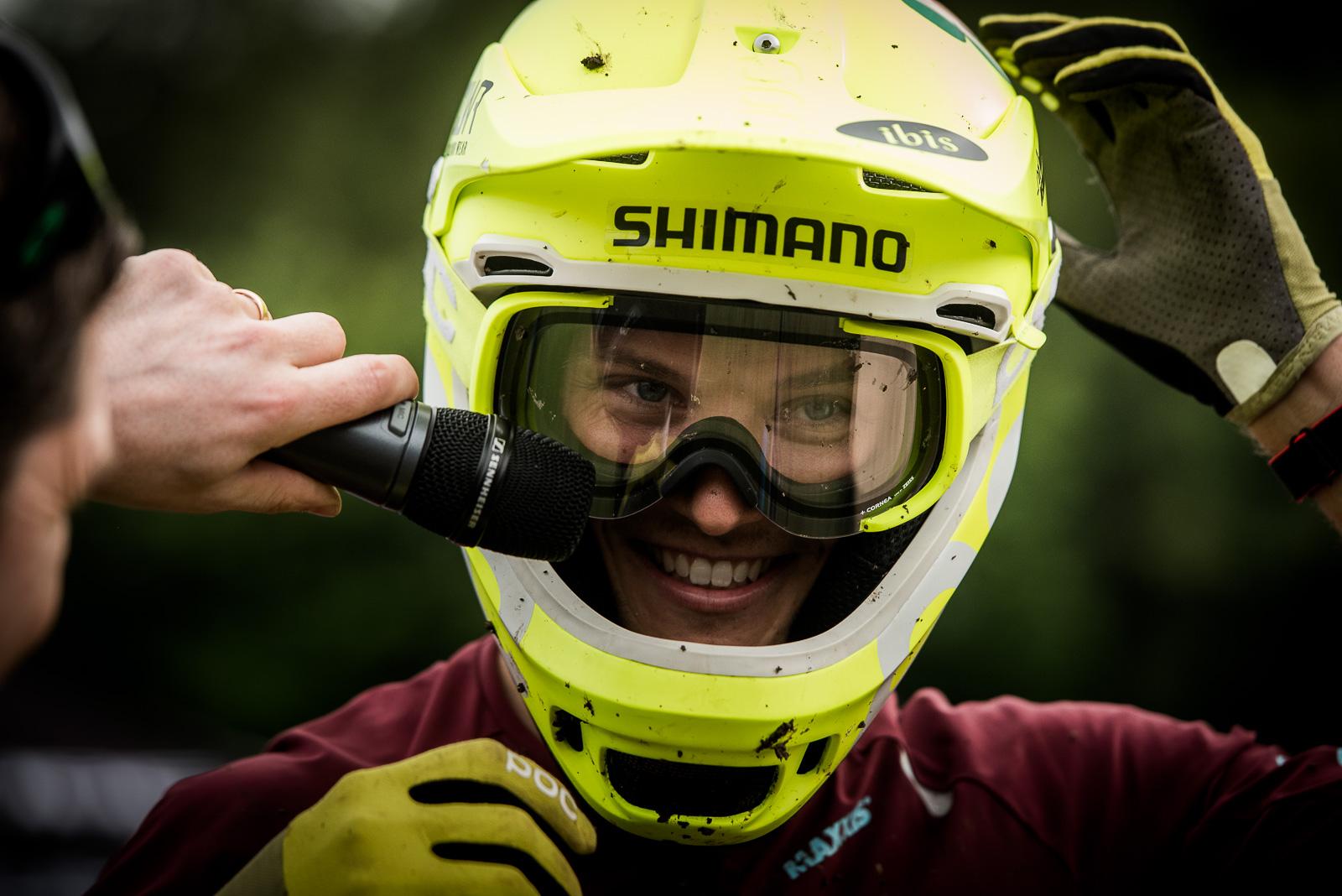 Ibis Mojo HD4: A person wearing a bright yellow helmet and goggles smiles while holding a microphone close to their face. The background is blurred, suggesting an outdoor setting. The individual is dressed in a maroon shirt with gloves on, and the helmet shows signs of wear with some dirt.