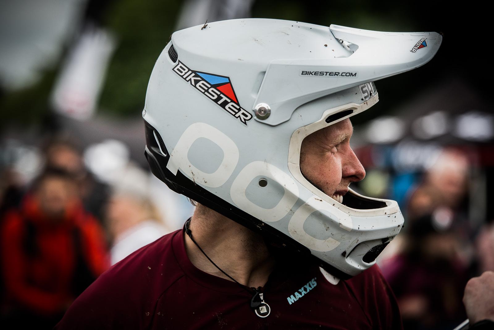 Ibis Mojo HD4: Side profile of a mountain biker wearing a white helmet with the logos "Bikester" and "POC". The biker is smiling and is dressed in a maroon shirt. The background features a blurred crowd and event setup, indicating an outdoor biking event. The helmet shows some dirt, suggesting recent use.