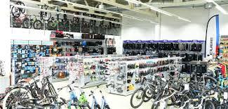 Interior view of a bicycle store displaying a variety of bicycles and cycling accessories. The space has bicycles lined up in the foreground, with racks showcasing helmets, tools, and gear along the walls. Additional bicycles are hung from the ceiling, maximizing display space. The store is well-lit and organized, creating an inviting shopping atmosphere for cycling enthusiasts.
