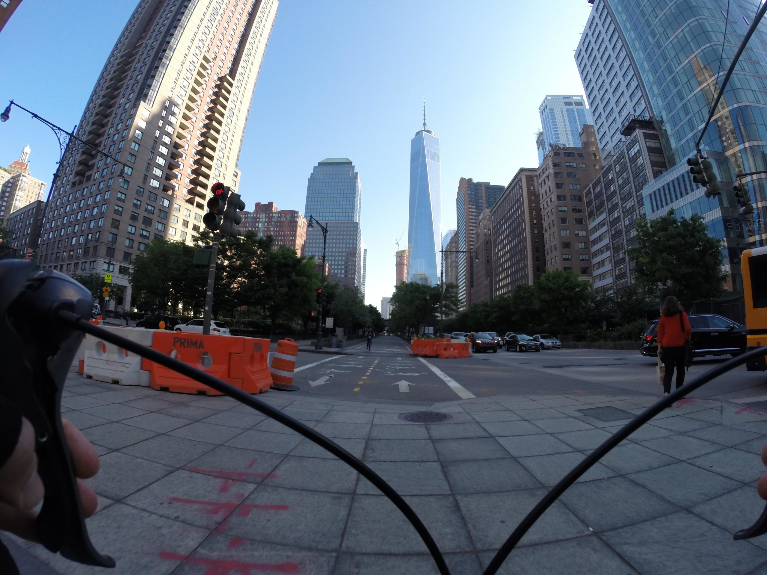 A view from the handlebars of a bicycle, looking down a city street lined with tall buildings. In the foreground, there is a green traffic light and construction barriers. In the distance, One World Trade Center is visible, rising above the cityscape under a clear blue sky. A person in a red jacket walks along the sidewalk, while cars are seen on the road. West Street Greenway mountain bike trail.
