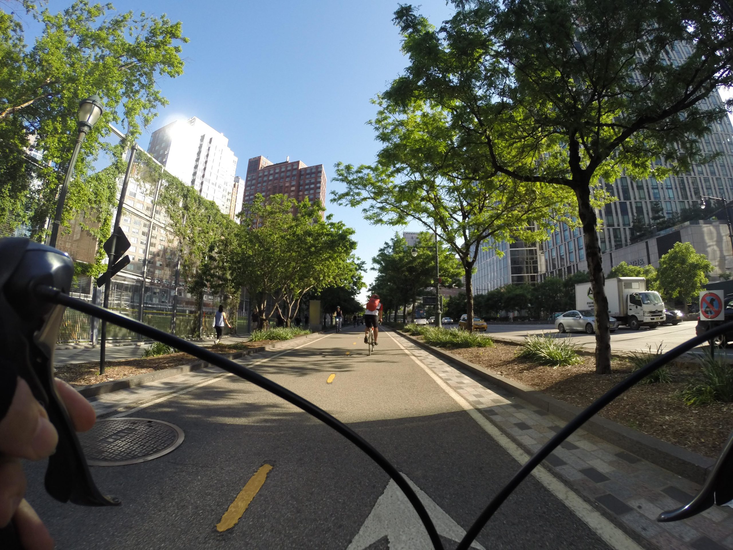A view from a cyclist's handlebars on a sunny day, showing a bike lane lined with trees and greenery. In the background, buildings reflect sunlight, and a few pedestrians and cyclists are visible along the path. The scene captures an urban environment with a mix of nature and city life. West Street Greenway mountain bike trail.
