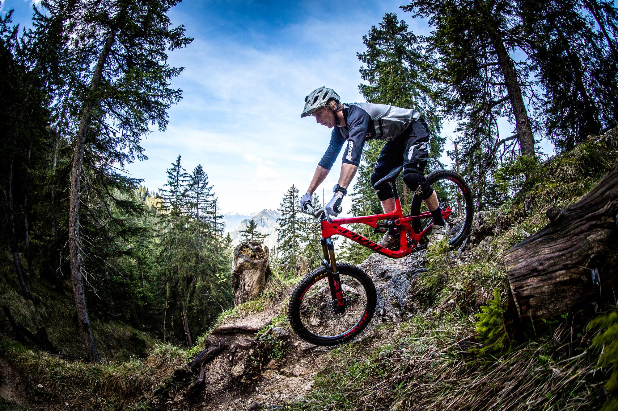 Pivot Mach 5.5: A mountain biker in protective gear navigates a rocky trail in a forested area, featuring tall trees and a clear blue sky in the background. The bike is red, highlighting the vibrant colors of the outdoor scene.