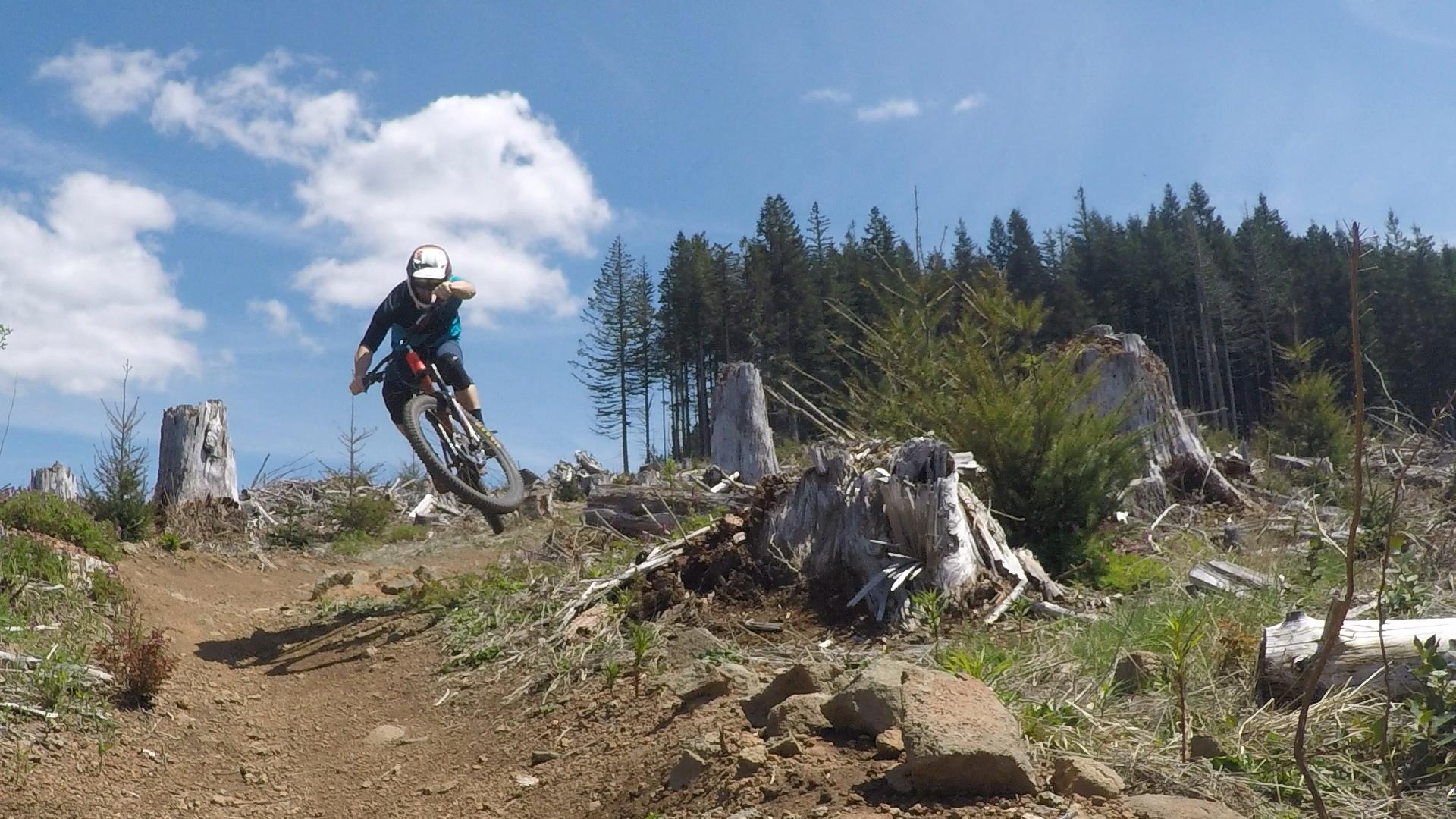 A mountain biker performing a jump on a dirt trail surrounded by tree stumps and lush greenery, under a bright blue sky with scattered clouds. Cold Creek mountain bike trail.
