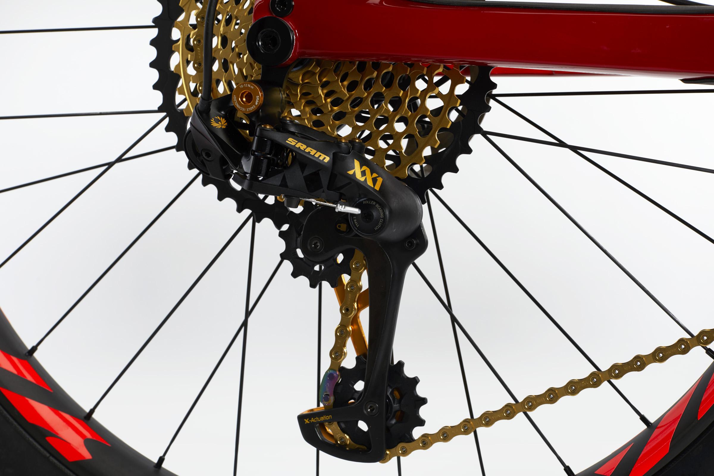 Ibis Mojo HD4: Image of a close-up view of a bicycle's rear derailleur, featuring a black SRAM XX1 derailleur, a gold cassette, and a gold chain. The background is minimalistic and white, focusing attention on the components.