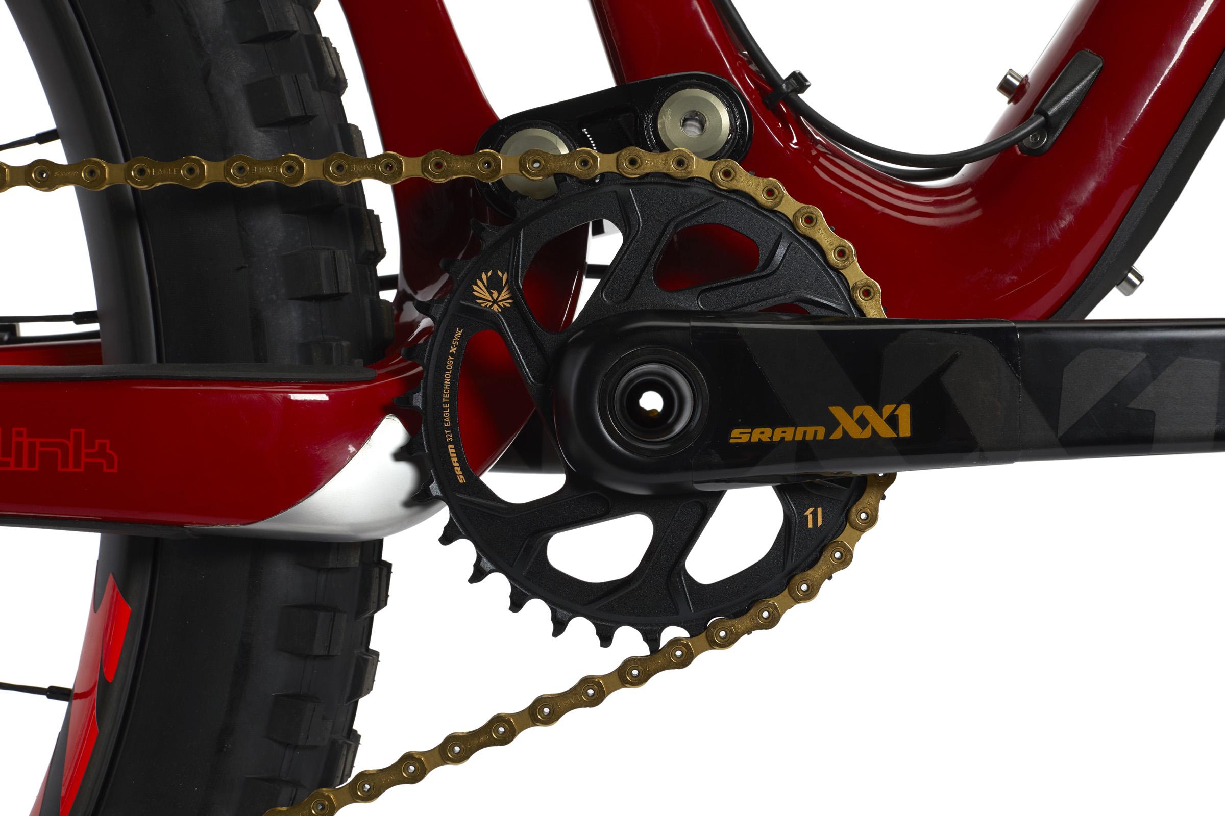 Ibis Mojo HD4: Close-up view of a mountain bike's drivetrain, featuring a black SRAM XX1 crankset with a gold chain, mounted on a vibrant red frame, showcasing the chainring and pedal assembly. The bike's robust tire is partially visible in the frame.