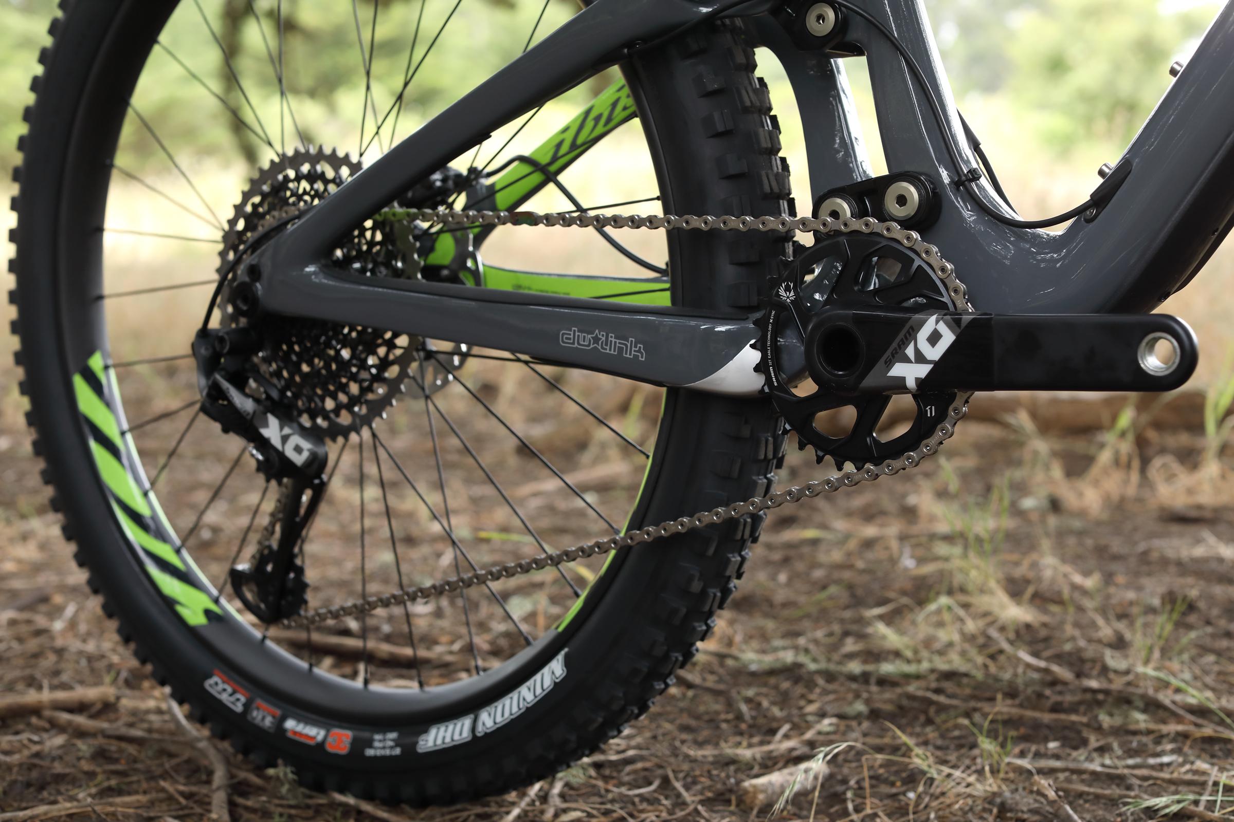 Ibis Mojo HD4: Close-up view of a mountain bike's drivetrain, featuring a black crankset with "SRAM XG" branding, a cassette, and chain. The bike's green and black tire is visible, along with the frame's gray and green color scheme. The background consists of dry grass and blurred greenery, indicating an outdoor setting.