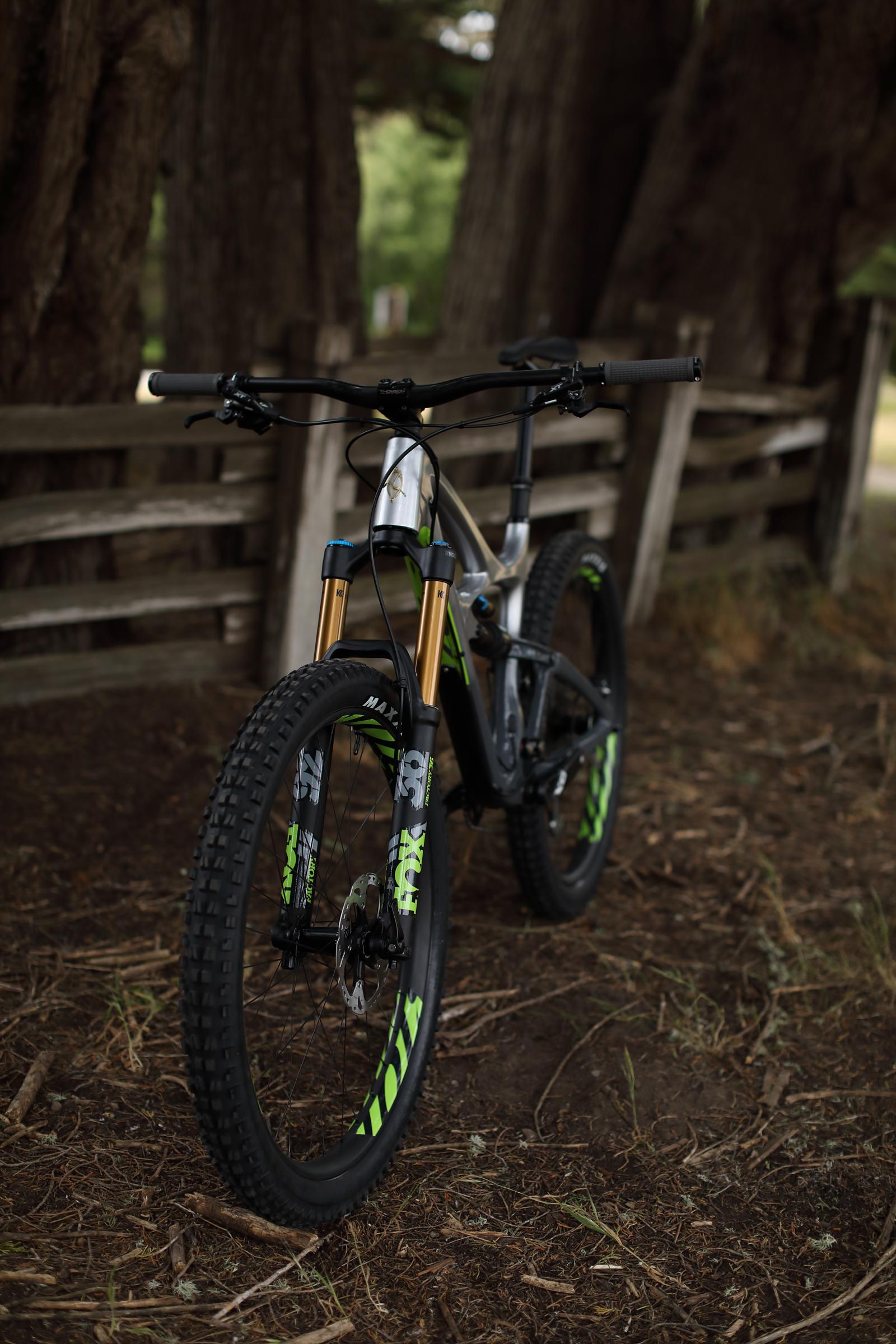 Ibis Mojo HD4: A mountain bike positioned on a dirt path surrounded by trees, featuring a sleek frame with a silver finish and green accents, along with prominent front forks and wide tires. A wooden fence is partially visible in the background.