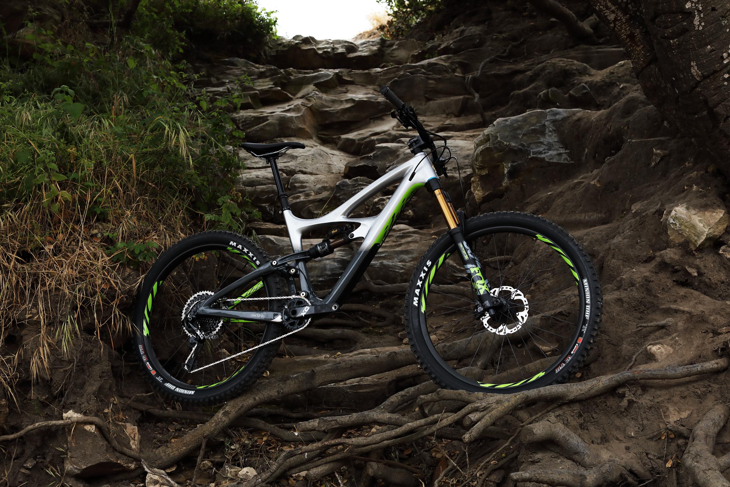 Ibis Mojo HD4: A mountain bike resting on rocky terrain surrounded by roots and vegetation, with a partially obscured path leading upward. The bike features a sleek silver and green frame, equipped with specialized tires designed for off-road riding.